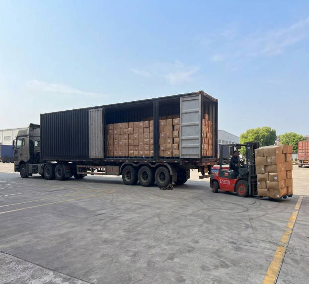 A full-scale logistics scene showing a large cargo truck being loaded with wholesale apparel boxes using a forklift at a professional distribution center.